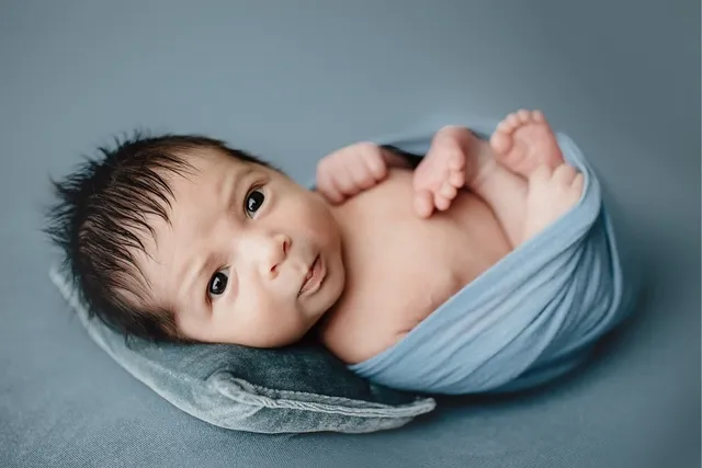 A wide-eyed newborn with dark hair wrapped in a light blue blanket on a soft gray backdrop