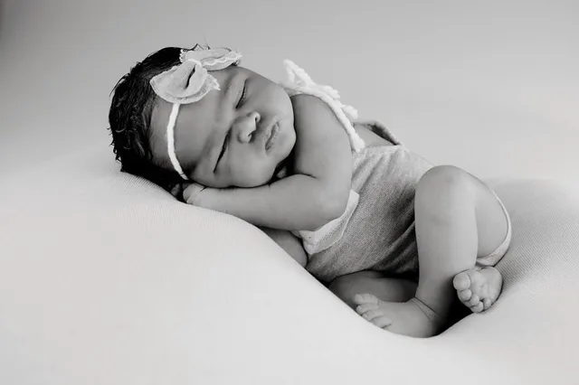 Black and white photo of a sleeping newborn curled on her side, wearing a bow headband