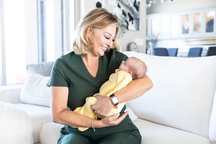 A Newborn Care Specialist in green scrubs smiling at a sleeping newborn