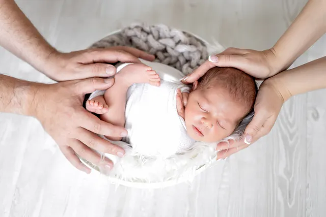 Two adults' hands resting on a sleeping newborn swaddled in white in a soft round basket