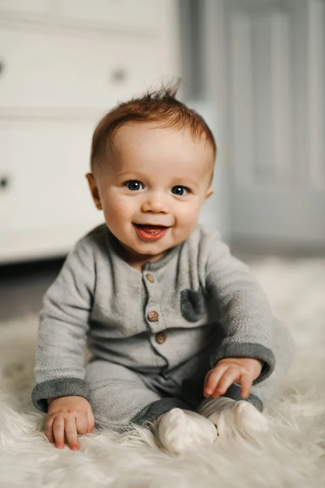 A smiling baby in a gray button-front outfit sitting on a white fluffy rug