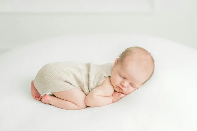 A sleeping newborn in a knit romper curled on a white cushion in a bright room