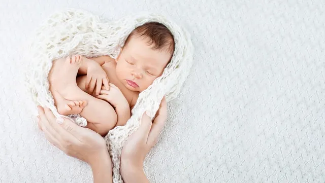 A sleeping newborn nestled in a heart-shaped white knit blanket cradled by an adult's hands