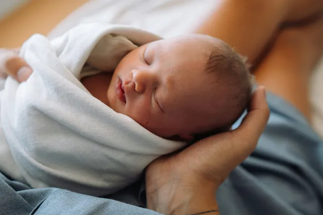 A sleeping newborn wrapped in a white blanket, cradled in an adult's hand