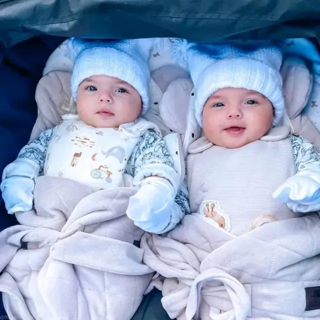 Two smiling infant twins in matching knit hats lying side by side in a stroller